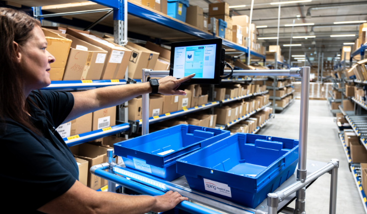 Préparation numérique des commandes dans l'entrepôt : un employé utilise un système Pick-by-Light avec écran tactile pour traiter les commandes. Deux caisses en plastique bleues sur un chariot, entourées d'étagères remplies de cartons d'expédition.